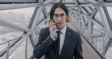 Modern cam koridorda yürüyen ve cep telefonuyla konuşan genç Asyalı işadamının pencerelerden metropole bakışını. İletişim ve iş konsepti.