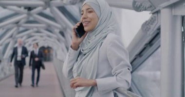 Orta Doğulu tesettürlü iş kadını cep telefonuyla konuşuyor. Modern cam salonda dikiliyor ve işadamları etrafta dolaşıyor. Cep telefonu iletişimi ve insan kavramı.