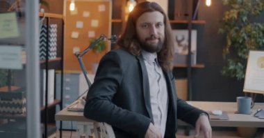 Takım elbiseli genç bir işadamının yavaş çekimde çekilmiş bir portresi. Masada oturuyor ve aydınlanmış ofiste kameraya bakıyor. İş ve kariyer kavramı.