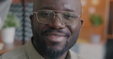 Kapalı alanda ofisin bulanık arka planına gülümseyen mutlu yüzüyle kameraya bakan Afrikalı Amerikalı iş adamının yavaş çekim portresi. İnsanlar ve olumlu duygu kavramı.