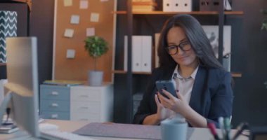 Akıllı telefon kullanan neşeli genç bir ofis çalışanı iş yerindeki masalara gülen akıllı telefon mesajlarını kontrol ediyor. Gadget ve iş konsepti.