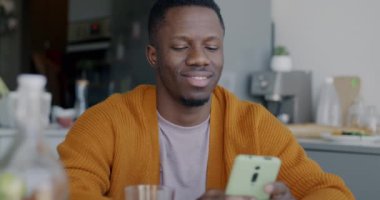 Joyful African American man using smartphone application touching mobile screen sitting at table in kitchen. Gadget and modern technology concept.