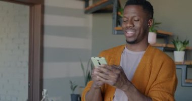 Cheerful African American man using smartphone messaging in social media holding gadget indoors in kitchen. People and internet communication concept.