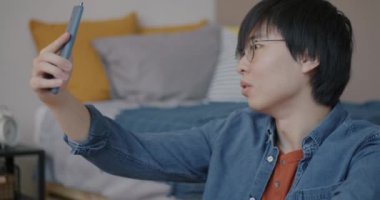 Neşeli Asyalı adam çevrimiçi video görüşmesi yapıyor. Yatak odasında akıllı telefon kullanarak sohbet ediyor. Sanal toplantı ve internet iletişim konsepti.