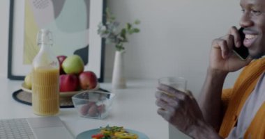 Neşeli Afrikalı Amerikalı adam cep telefonuyla konuşuyor ve evdeki mutfak masasında portakal suyu içiyor. İletişim ve gençlik yaşam tarzı kavramı.