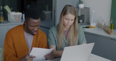 Beyaz kadın ve Afro-Amerikalı adam evdeki mutfak masasında oturmuş laptoplarını daktilo edip kağıt okuyorlar. Aile ve modern teknoloji kavramı.