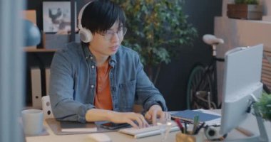 Bilgisayarda çalışan Asyalı işadamı ofis masasında kulaklıkla müzik dinliyor ve daktilo ediyor. Gençlik kültürü ve modern yaşam tarzı kavramı.