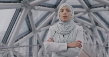 Modern cam salonda kollarını kavuşturup kameraya bakan, tesettürlü, kendinden emin Orta Doğulu iş kadınının yavaş çekimde portresi. Müslüman iş kadını ve girişimci konsepti.