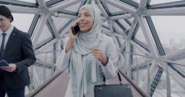 Orta Doğulu tesettürlü bir iş kadını modern cam koridorda yürüyor cep telefonuyla konuşuyor ve pencerelerden şehri izliyor. İş ve iletişim kavramı.