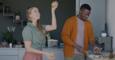 Afro-Amerikalı erkek salata pişirirken, beyaz kadın da evde gülerek meyvelerle hokkabazlık yapıyor. Beslenme ve aile yaşam tarzı konsepti.