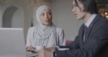 Orta Doğulu tesettürlü iş kadını Asyalı erkek partnerle konuşuyor ve kafede dizüstü bilgisayar ekranına bakıyor. İş ortaklığı ve teknoloji kavramı.