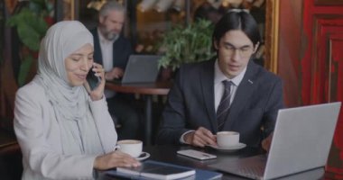 Orta Doğulu iş kadını cep telefonuyla konuşuyor ve kafedeki masada dizüstü bilgisayarını kullanarak Asyalı iş arkadaşına bilgi paylaşıyor. Profesyonel iletişim ve iş konsepti.