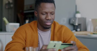 Afro-Amerikalı bir adam evdeki mutfak masasında oturan akıllı telefonuyla alışveriş yapıyor ve kredi kartıyla ödüyor. Tüketim ve bankacılık kavramı.