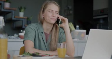 Neşeli genç bayan cep telefonuyla konuşuyor ve evdeki mutfak masasında yemeğin tadını çıkarıyor. Cep telefonu iletişimi ve yemek zamanı kavramı