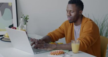 Afrikalı Amerikalı adam bilgisayarla çalışıyor ve evdeki mutfak masasında öğle yemeğinde sandviç yiyor. Serbest meslek ve beslenme konsepti.