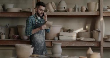 Küçük işletme sahibi cep telefonuyla konuşuyor raflardaki tencereleri kontrol ediyor çömlekçilik atölyesi geliştiriyor. İletişim ve geleneksel sanat stüdyosu konsepti.