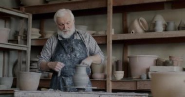 Kıdemli sanatçı tekerlek fırlatmaktan seramik kap hazırlıyor ve atölyede rafa koyuyor. Yetenekli çömlekçi küçük işletmeler kurmaktan memnun..