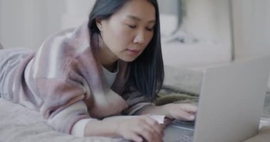 Asyalı serbest çalışan kadının dizüstü bilgisayar kullanması ve yatak odasında yatarak defter planlama projesi yazması. Serbest meslek ve insanların konsepti.