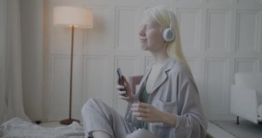 Pijamalı mutlu albino kızın kulaklıkla müzik dinlemesi ve lüks yatak odasında yatakta akıllı telefon tutarak dans etmesi. Gençlik kültürü ve eğlence konsepti.