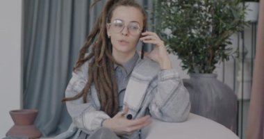 Rasta saçlı güzel bir bayan cep telefonuyla konuşuyor oturma odasındaki lüks bir dairede koltukta oturuyor. İletişim ve cep telefonu kavramı.