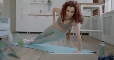 Spor kıyafetli çekici genç bir kadın evde egzersiz yapıyor ve bilgisayar ekranında video eğitimi izliyor. Sağlıklı yaşam tarzı ve modern teknoloji kavramı.