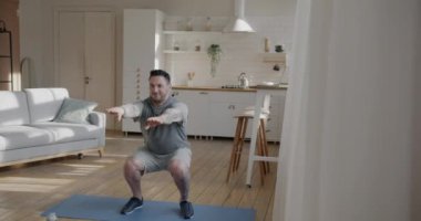 Spor giyimli formalı genç adam evde spor antrenmanlarına konsantre olarak çömelme egzersizleri yapıyor. Spor ve aktif yaşam tarzı kavramı.