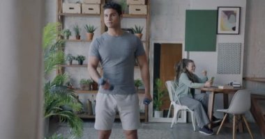 Orta Doğulu adam dambıllarıyla egzersiz yaparken neşeli Kafkas kız arkadaşı dairede akıllı telefonuyla online video görüşmesi yapıyor. Spor ve modern yaşam tarzı konsepti.