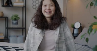 Neşeli Asyalı bir kızın evde oturup kameraya bakarak mutluluğunu ifade ederek gülüşünün yavaş çekim portresi. İnsanlar ve iyi ruh hali kavramı.