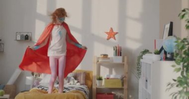 Süper kahraman kostümlü, yatakta zıplayıp evde komik oyunlar oynayan hiperaktif bir kız. Çocukluk ve eğlence kavramı.