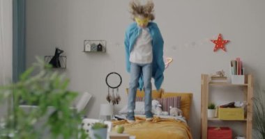 Süper kahraman kostümü giymiş, yatakta zıplayıp yatak odasındaki boş aktivitelerin keyfini çıkaran mutlu bir çocuğun ağır çekimde portresi. Çocukluk ve eğlence kavramı.