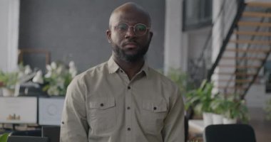 Ofiste durmuş, ciddi bir yüzle kameraya bakan Afro-Amerikan genç bir adamın yavaş çekim portresi. Çalışan ve mesleki meslek kavramı.