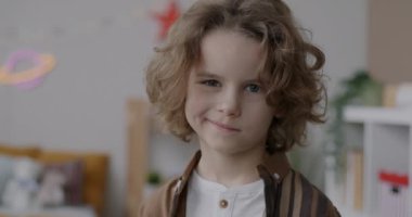 Evin arka planında komik bir yüz ifadesiyle kameraya bakan sevimli küçük çocuğun yavaş çekim portresi. Çocukluk ve duygu kavramı.