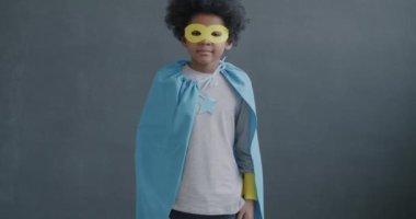 Süper kahraman maskeli ve pelerinli neşeli Afro-Amerikan çocuğun gri arka planda durup kameraya gülümseyen portresi.
