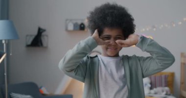 Evde kameraya bakarak komik maske hareketleri yapan sevimli Afrikalı Amerikalı çocuğun yavaş çekim portresi. Çocukluk ve eğlence kavramı.