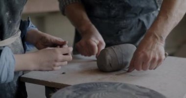 Büyükbaba atölyedeki çocuğa ders verirken yetişkin ve çocuğun kille çalıştığı yakın çekim. Çömlekçilik ve aile mesleği.
