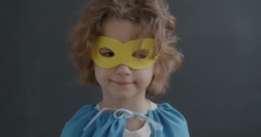 Sarı saçlı, maske takan, süper kahraman kılığına girmiş, gri arka planda kameraya gülümseyen tatlı bir çocuğun yakın plan portresi. Çocukluk ve eğlence kavramı.