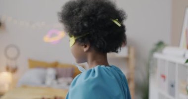 Süper kahraman maskeli komik Afro-Amerikan çocuğun yavaş çekim portresi evde eğlenirken gülümseyen kameraya dönüşüyor. Çocukluk ve eğlence kavramı.