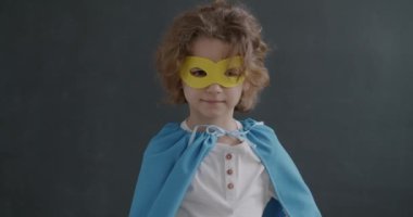 Süper kahraman maskeli ve pelerinli, kameraya bakıp gülümseyen sevimli sarışın çocuğun yavaş çekimde portresi. Çocukluk ve oyun konsepti.