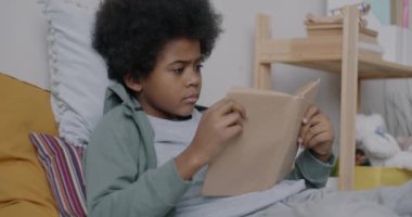 Zeki Afro-Amerikan çocuk yatakta yatan ilginç bir kitap okuyor dairede yatak odasında vakit geçiriyor. Çocukluk ve eğitim kavramı.