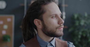 Rasta saçlı genç bir adamın yakın çekim yavaş portresi kameraya dönüyor ve içeride yaratıcı ofiste gülümsüyor. İnsanlar ve bireysellik kavramı.