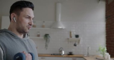 Dambıllarla egzersiz yapan kaslı bir adam dairede çalışıyor ve kablosuz kulaklıkla müzik dinliyor. Aktif yaşam tarzı ve vücut geliştirme kavramı.