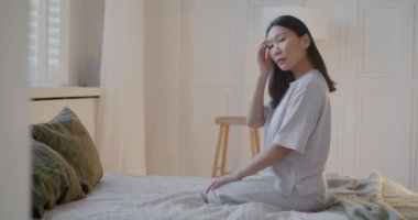 Pijamalı, şık bir yatak odasında yatağında kameraya bakan güzel Asyalı kadının yavaş çekimde portresi. Ev ve insanlar kavramı.