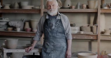 Arka planda seramik dolu raflarla çömlekçilik atölyesinde tek başına duran çamurlu önlüklü yaşlı bir adamın yavaş çekim portresi. Geleneksel sanatlar ve yaratıcı meslek kavramı.
