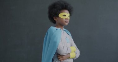 Ağır çekimde süper kahraman kostümü giymiş, gri arka planda kolları çapraz duran ve ciddi bir yüzle kameraya bakan küçük Afrikalı Amerikalı çocuğun portresi.