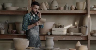 Küçük işletme sahibi cep telefonuyla konuşuyor raflardaki tencereleri kontrol ediyor çömlekçilik atölyesi geliştiriyor. İletişim ve geleneksel sanat stüdyosu konsepti.