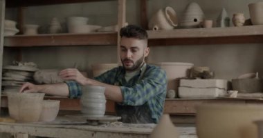 İş yerindeki yaratıcı aktivitelere yoğunlaşmış çömlek çarkı kullanarak vazo yapan genç adamın yavaş çekimde portresi. İnsanlar ve küçük işletme kavramı.