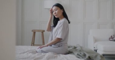 Lüks yatak odasında rahat bir yatakta pijama giyen ve kameraya bakan çekici Asyalı kadının yavaş çekimde portresi. İnsanlar ve iç konsept.