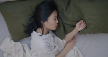Asyalı bir kadının yatakta uyuyup yastıkta dinlenmesinin ve yatak odasında uyuyan rahat çarşafların keyfini çıkarmasının en iyi görüntüsü. Dinlenme ve yatak konsepti.