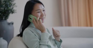 Lüks apartmandaki koltukta oturan genç Asyalı kadın cep telefonuyla konuşuyor. Cep telefonu iletişimi ve kablosuz teknoloji kavramı.