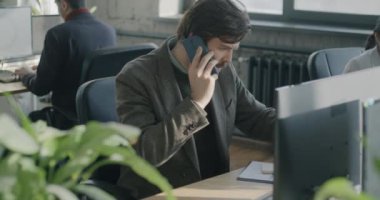 Genç ofis çalışanı cep telefonuyla konuşuyor ve ortak iş yerinde not defterine bilgi yazıyor. İş iletişimi ve cep telefonu arama kavramı.